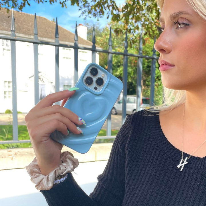 A woman with long blonde hair holds an iPhone adorned with Coconut Lane's Melting Heart Phone Case in Baby Blue. She's wearing a black sweater and has a hair scrunchie on her wrist. A fence and a white house are visible in the background.