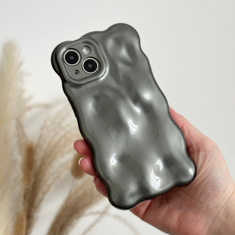 A hand holding an iPhone encased in a 3D Pearl Phone Case - Grey from Coconut Lane. The case features an extra protective, metallic, wavy, and textured design with an irregular surface that gives it a shiny, reflective appearance. In the blurred background, some dried decorative grasses can be seen.