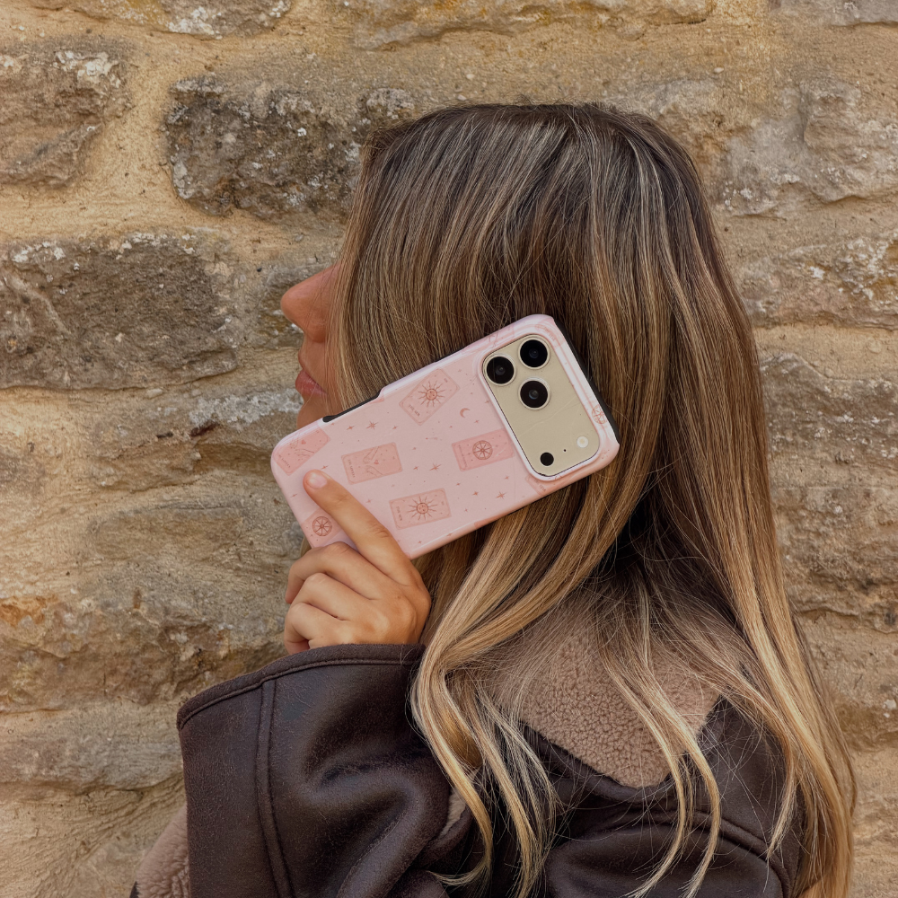 Woman holding a phone up to her ear, showing a light pale pink phone case with tarot cards and tiny stars. The woman is wearing a brown aviator jacket. 