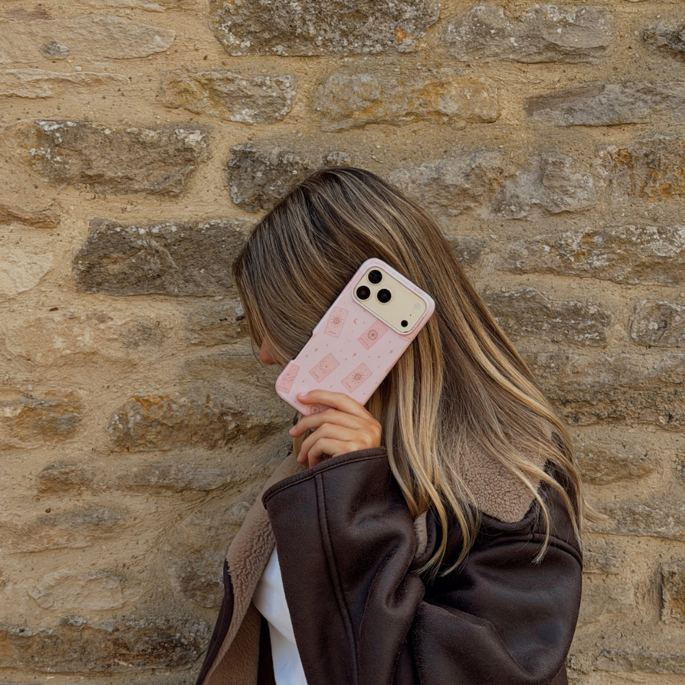 Woman holding a phone up to her ear, showing a light pale pink phone case with tarot cards and tiny stars. The woman is wearing a brown aviator jacket. 
