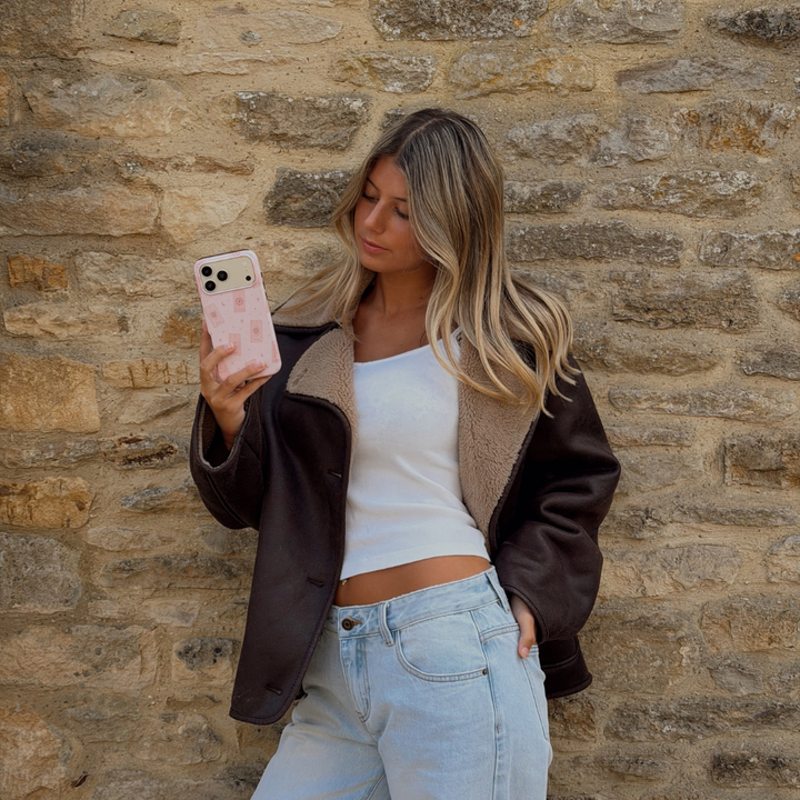 Woman holding a phone up taking a photo, showing a light pale pink phone case with tarot cards and tiny stars. The woman is wearing a brown aviator jacket and light blue jeans.