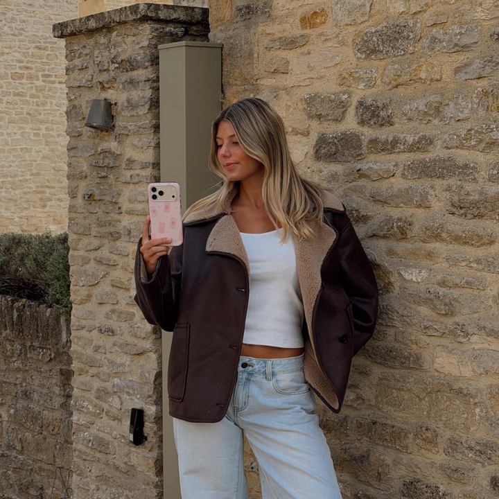 Woman holding a phone up taking a photo, showing a light pale pink phone case with tarot cards and tiny stars. The woman is wearing a brown aviator jacket and light blue jeans.