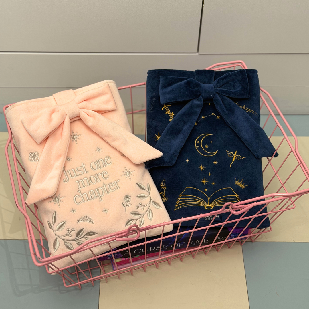 Book Sleeve made from soft blush pink velvet with grey embroidered floral details, stars and 'Just One More Chapter' lettering, finished with a large pink velvet bow and Coconut Lane logo, placed in a pink shopping basket.