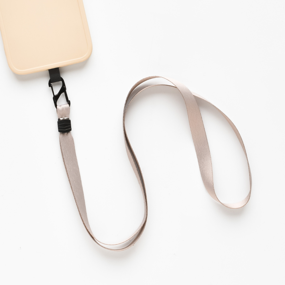 The Universal Phone Lanyard Patch Bundle by Coconut Lane, featuring a beige strap and tan lanyard, is displayed on a white surface. Designed specifically as a phone accessory, it includes a black plastic buckle and clip that are partially visible near the top left corner of the image. The strap itself is coiled in a loose loop.