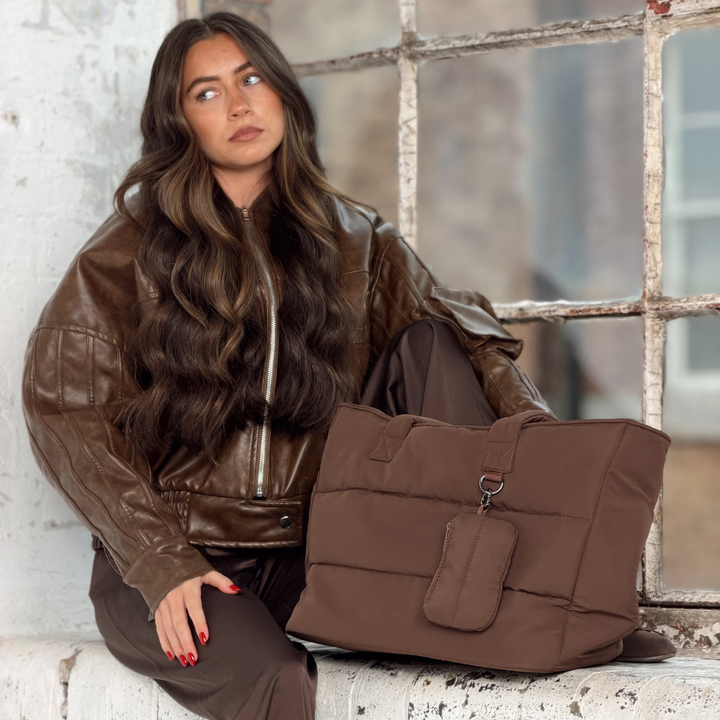 Woman with long brunette hair wearing a brown leather jacket. Next to her is the Coconut Lane Main Carry Choc Puffer bag with the matching Chocolate brown mini keyring bag with zip fastening, silver metal heart shaped clip attached to the outside D ring. 