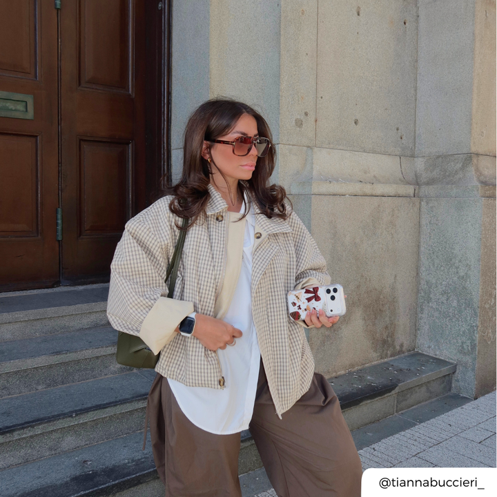A woman stands on stone steps wearing a beige and white gingham cropped jacket over a white top and brown trousers, carrying a green shoulder bag and holding a phone in a clear phone case decorated with a red bow, cherries, a martini glass, a strawberry dipped in chocolate, and small star accents. 