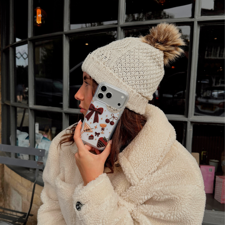 A woman wearing cream knit gloves and a cream teddy coat, holds up a clear phone case decorated with a red bow, cherries, a martini glass, a strawberry dipped in chocolate, and small star accents. The background shows a cafe setting.