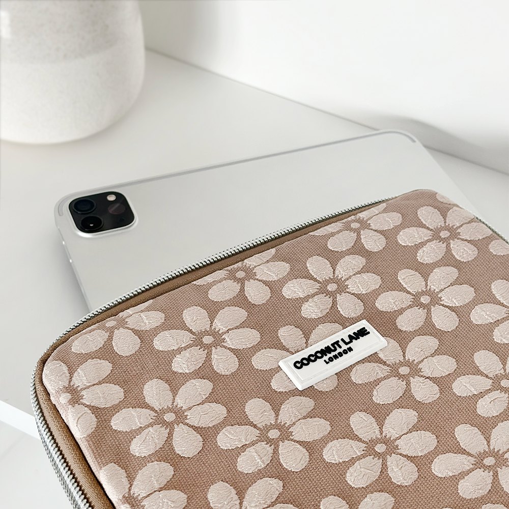 A Mocha Flower iPad Sleeve, featuring a beige color and embroidered white floral pattern, sits partially zipped on a white surface. A silver laptop and a modern white vase are visible in the background. The label on the sleeve reads, "Coconut Lane.