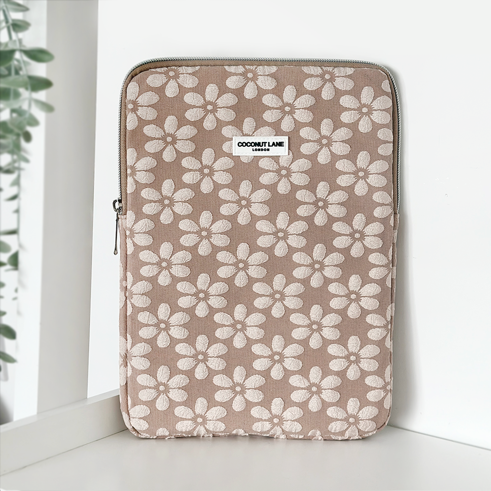 The Mocha Flower iPad Sleeve by Coconut Lane, featuring a beige background with white flowers scattered across its surface, is placed on a white surface. It has a zip closure along the top edge and a small "Coconut Lane" label near the top center. A green potted plant is partially visible to the left.