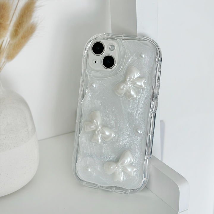 A **Coconut Lane** Pearls & Bows Phone Case, adorned with white textured bows and pearl-like accents, rests on a white shelf. Nearby, a white vase filled with dried wheat stalks adds a decorative touch to the background.