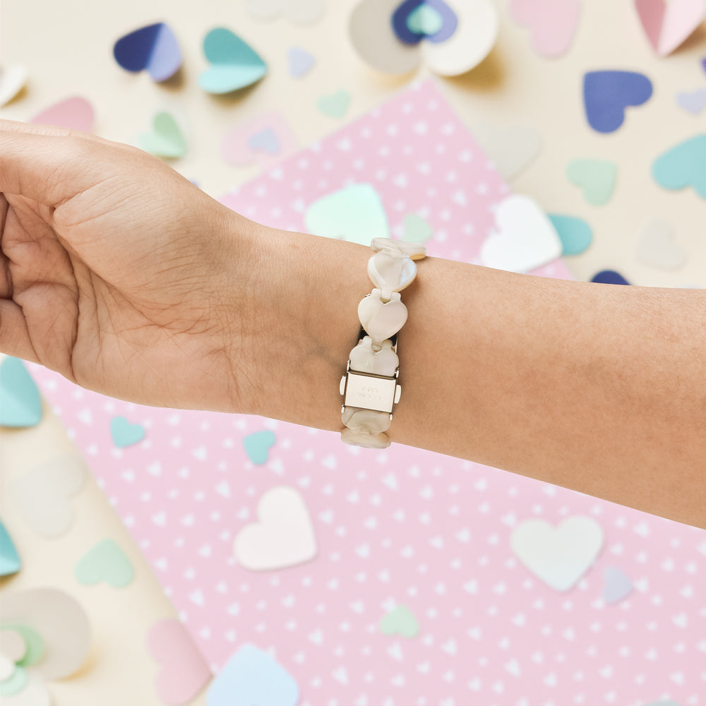 A wrist is shown wearing the Luxe Hearts Apple Watch Strap - Rose Quartz by Coconut Lane, adorned with white heart-shaped beads. The background features scattered colorful paper hearts in shades of blue, pink, and cream, set against a pink polka-dotted paper beneath the arm.
