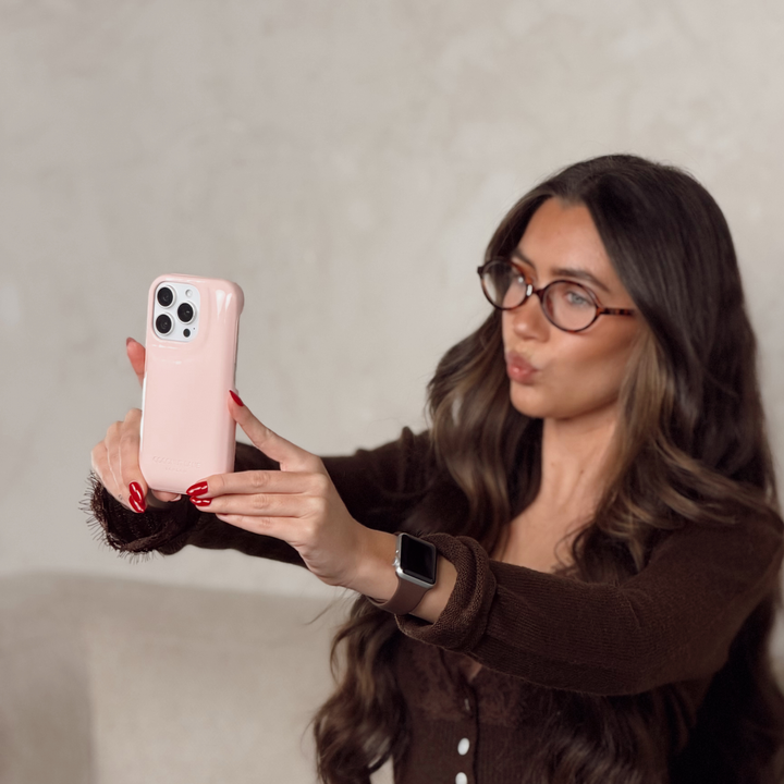 Woman in a brown cardigan with lace detail, is holding an iphone in a glossy pastel pink Coconut Lane puffer phone case with embossed logo taking a selfie. 
On her wrist is a Coconut Lane NAKD Chocolate brown apple watch strap.
