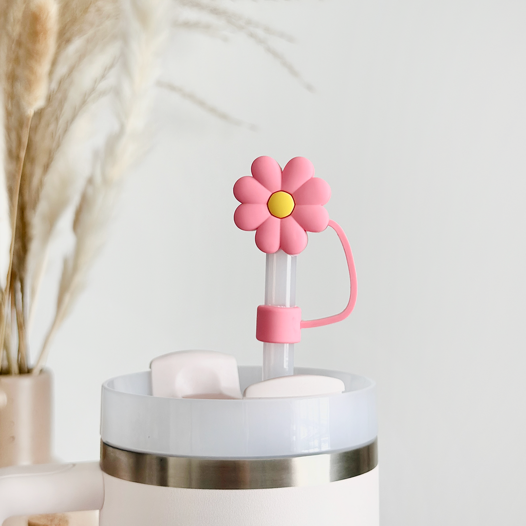 A tumbler with a lid is filled with white ice cubes. The lid features a protruding straw topped with a Pink & Yellow Daisy Straw Cover from Coconut Lane. In the background, there is a beige vase with dried pampas grass. The setting is minimalistic, accented by the simple white background and silicone details on the straw.