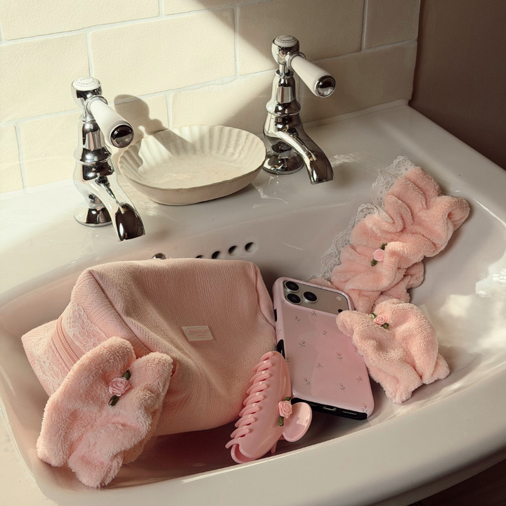 A collection of soft pink bath accessories arranged in a white sink, including plush bath towels, a claw hair clip, a phone case, and a scrunchie, with chrome faucets and a tiled wall in the background.