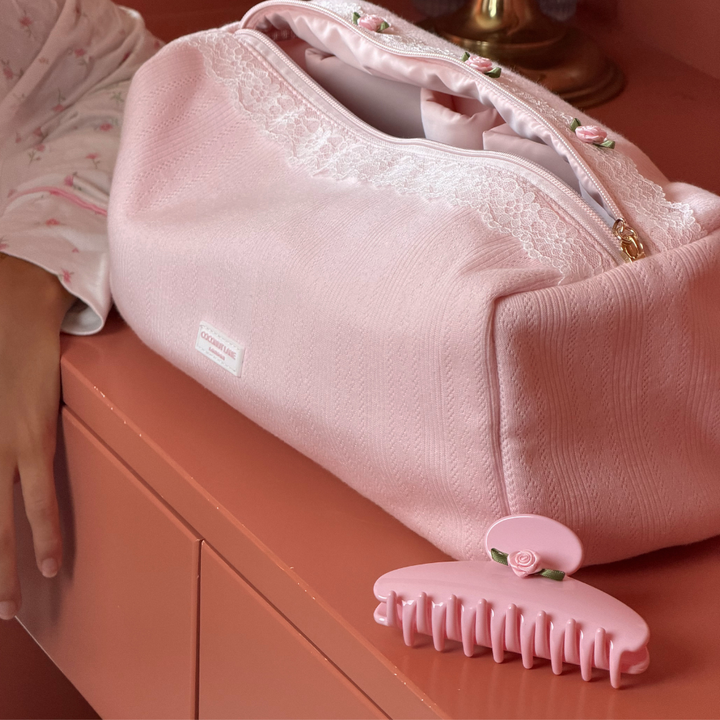 A pink textured fabric zippered bag with white lace trim and small floral embroidery, resting on a peach-colored surface next to a matching pink hair claw clip with a small rose flower detail. 