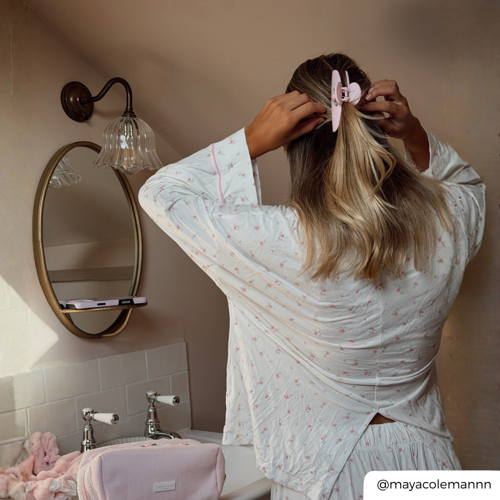 A woman with blonde-highlighted hair seen from behind, wearing a white long-sleeve pajama top with a tiny pink floral pattern and matching shorts. She is adjusting her hair with a light pink claw clip with a small rose detail on it, in a bathroom setting with a sink, mirror.