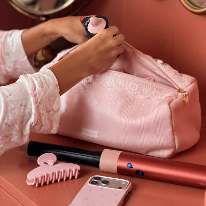 A pink hair tool bag with white lace trim and a gold zipper, resting on a peach-colored cabinet next to a person's hand. A matching pink hair claw clip is in the foreground.