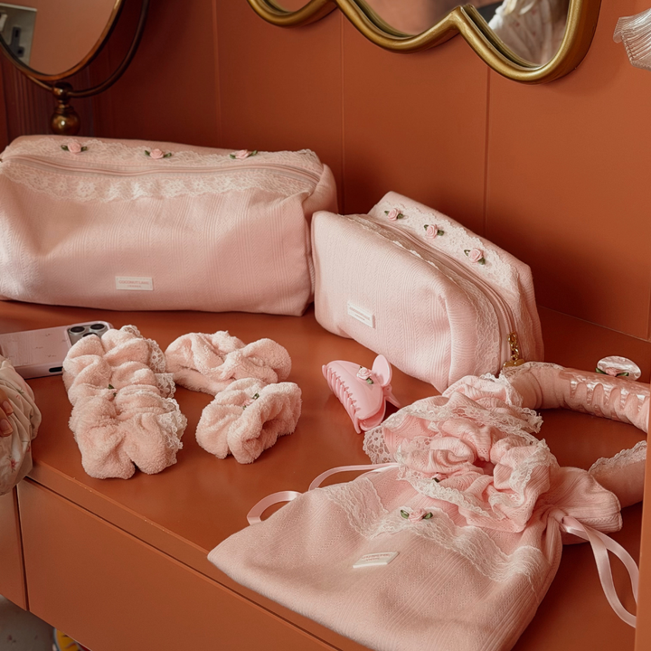 A coordinated set of baby pink vanity accessories arranged on a warm terracotta vanity surface, including a makeup bag and a hair tool bag, with a pink hair clip, matching headband and wristband set and a and a lace-trimmed drawstring pouch.