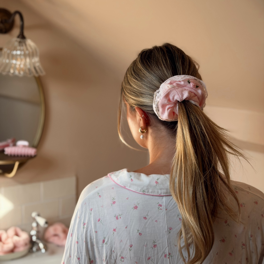 A woman viewed from behind wearing a light pink ruffled scrunchie with small embroidered rose, holding her blonde ponytail. She is wearing a white floral pajama top in a warm-toned bathroom setting.