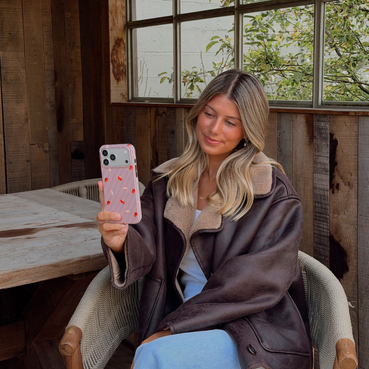 Woman wearing a brown aviator jacket holding up an iphone taking a selfie. The phone is in an autumnal case, with brown vertical stripes, and small prints of pumpkin spice lattes and sweet treats.