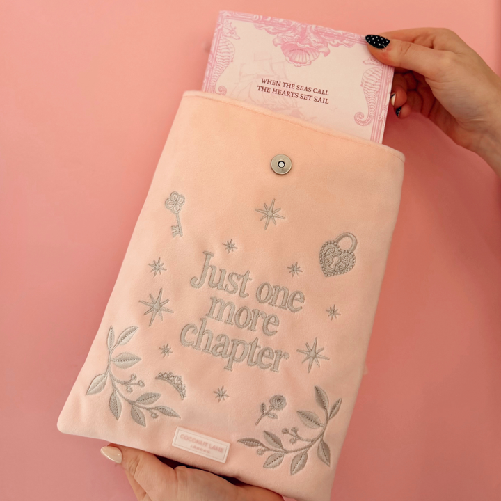 Book Sleeve made from soft blush pink velvet with grey embroidered floral details, stars and 'Just One More Chapter' lettering, finished with a large pink velvet bow and Coconut Lane logo, 
