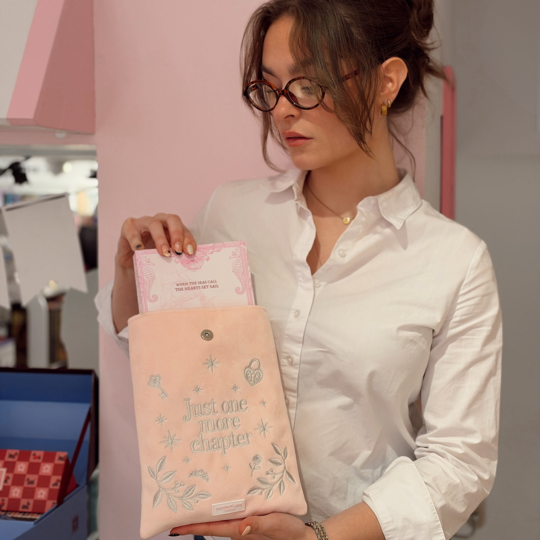 Woman in bookstore, holding a Book Sleeve made from soft blush pink velvet with grey embroidered floral details, stars and 'Just One More Chapter' lettering, finished with a large pink velvet bow and Coconut Lane logo, 