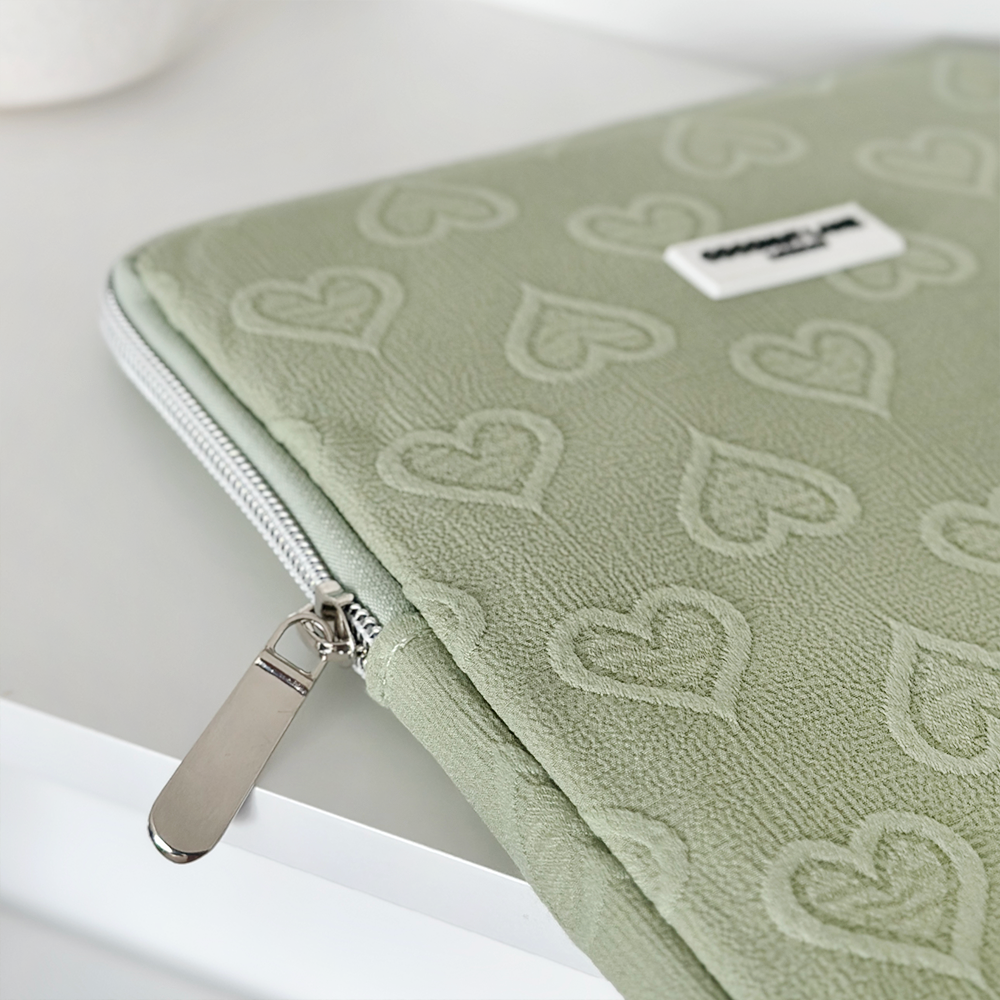 A close-up of the Sage Heart iPad Sleeve by Coconut Lane, featuring a light green fabric with a silver zipper. The exterior is embossed with a heart pattern, making it ideal for an 11" iPad. There is a small rectangular tag with text sewn near the zipper. The pouch rests on a white surface.
