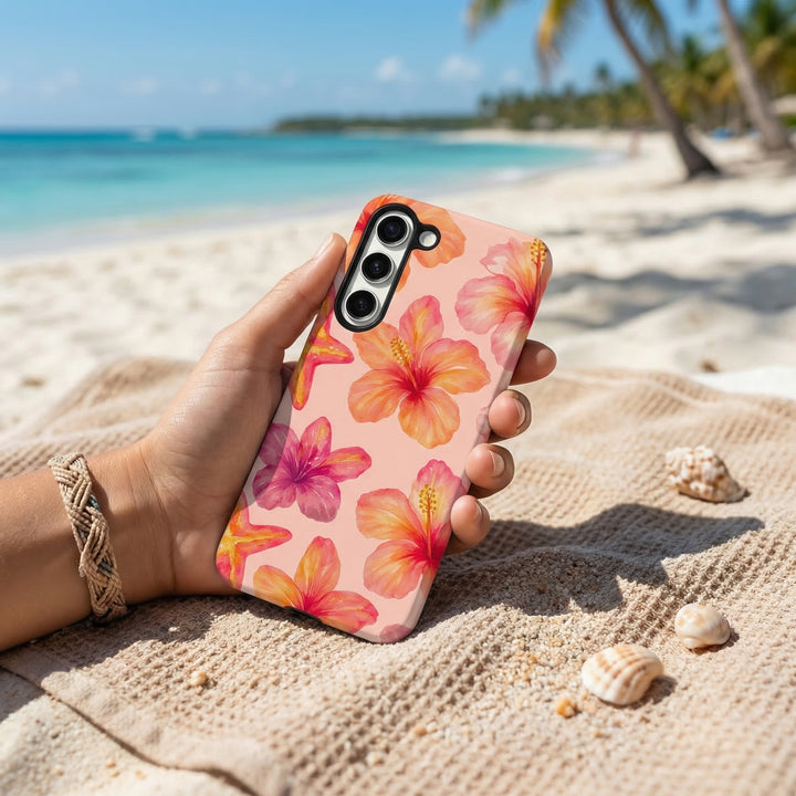Hand holding a pink hibiscus Samsung Tough Phone Case on a sandy beach with seashells and palm trees in the background.