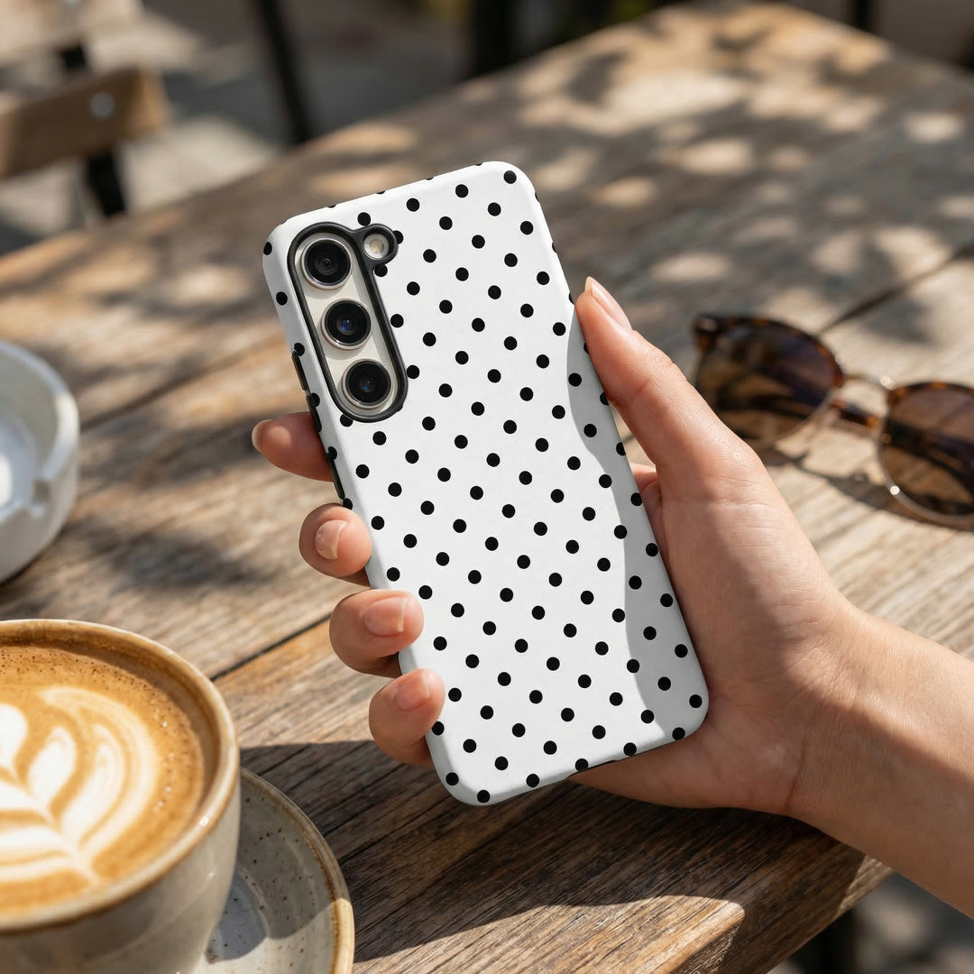 Hand holding white polka dot Samsung phone case by Coconut Lane beside sunglasses and a latte on a table.