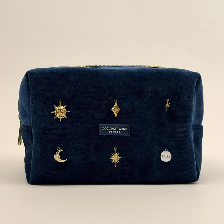 Stardust After Dark makeup bag. Dark navy blue velvet material with gold zip, and sewn-on gold celestial-themed charms including a sun, moon, star, diamond shape and 11:11 charm.  