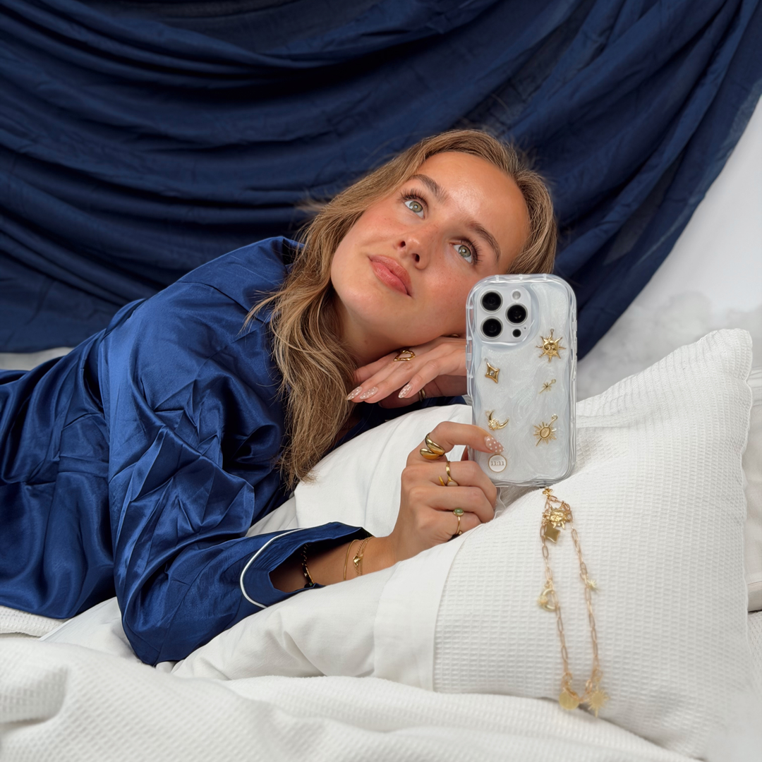 Woman wearing satin navy pyjamas laying in a bed with white bedding, She is holding a Stardust after dark charm phone case. A pearl base with luxe gold celestial charms including a moon, sun, star, diamond shape and 11:11 charm. Attached to the phone case is a matching gold chain phone strap. 