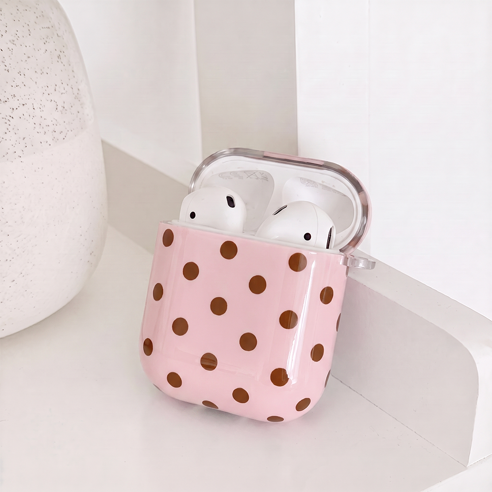 Showing the inside of a glossy pink case for AirPods with brown polka dots and a small clear clip loop on the side. It sits on a white shelf beside a white speckled vase.