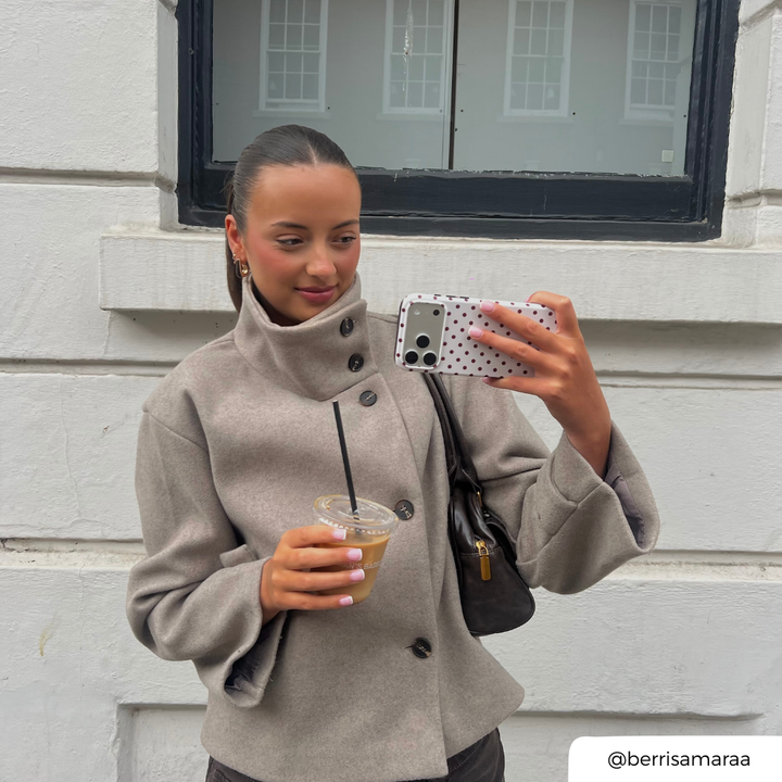 A woman takes a selfie outdoors against a white brick wall, wearing a light taupe high-neck button-up coat and holding an iced coffee. The phone case is cream with brown polka dots. 