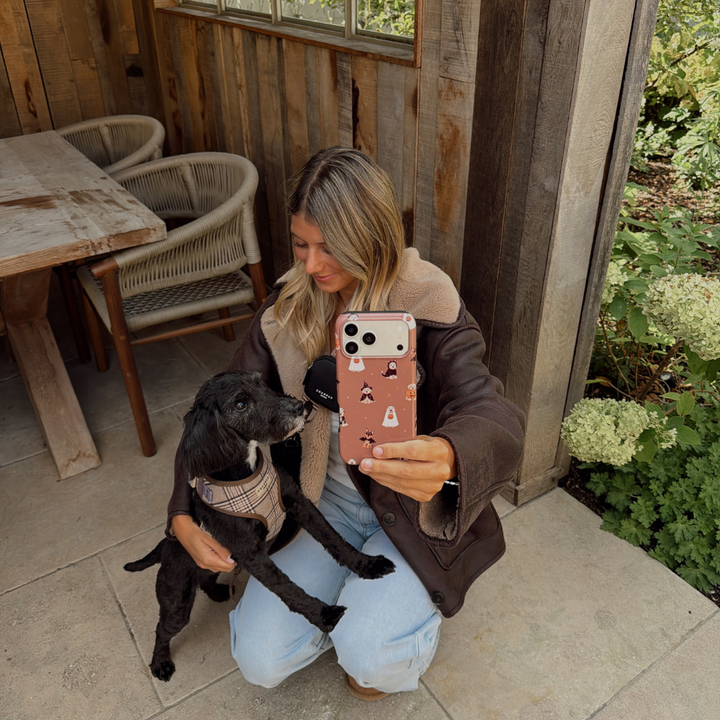 Woman in a brown aviator jacket, holding a phone case with dogs wearing spooky outfits on a brown background, whilst kneeling on the ground stroking a black dog. 