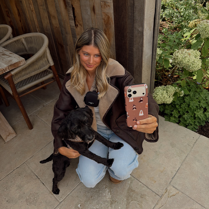 Woman in a brown aviator jacket, holding a phone case with dogs wearing spooky outfits on a brown background, whilst kneeling on the ground stroking a black dog. 