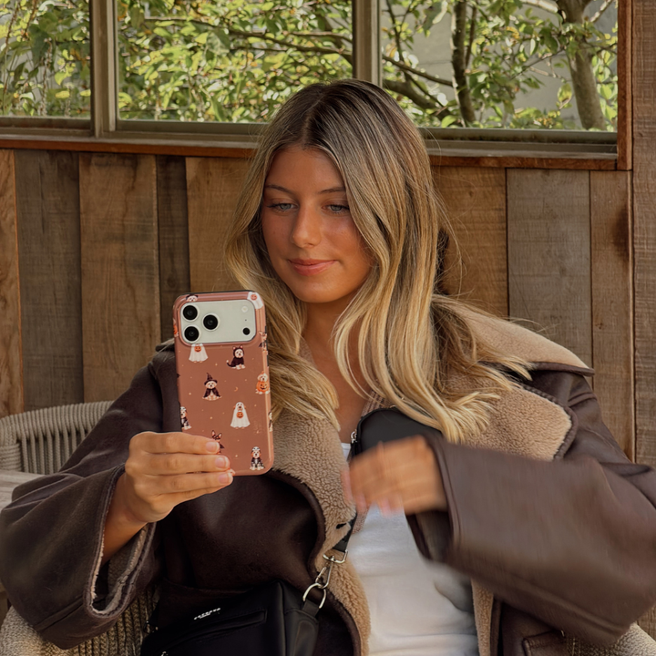 Woman in a brown aviator jacket, holding a phone case with dogs wearing spooky outfits on a brown background.