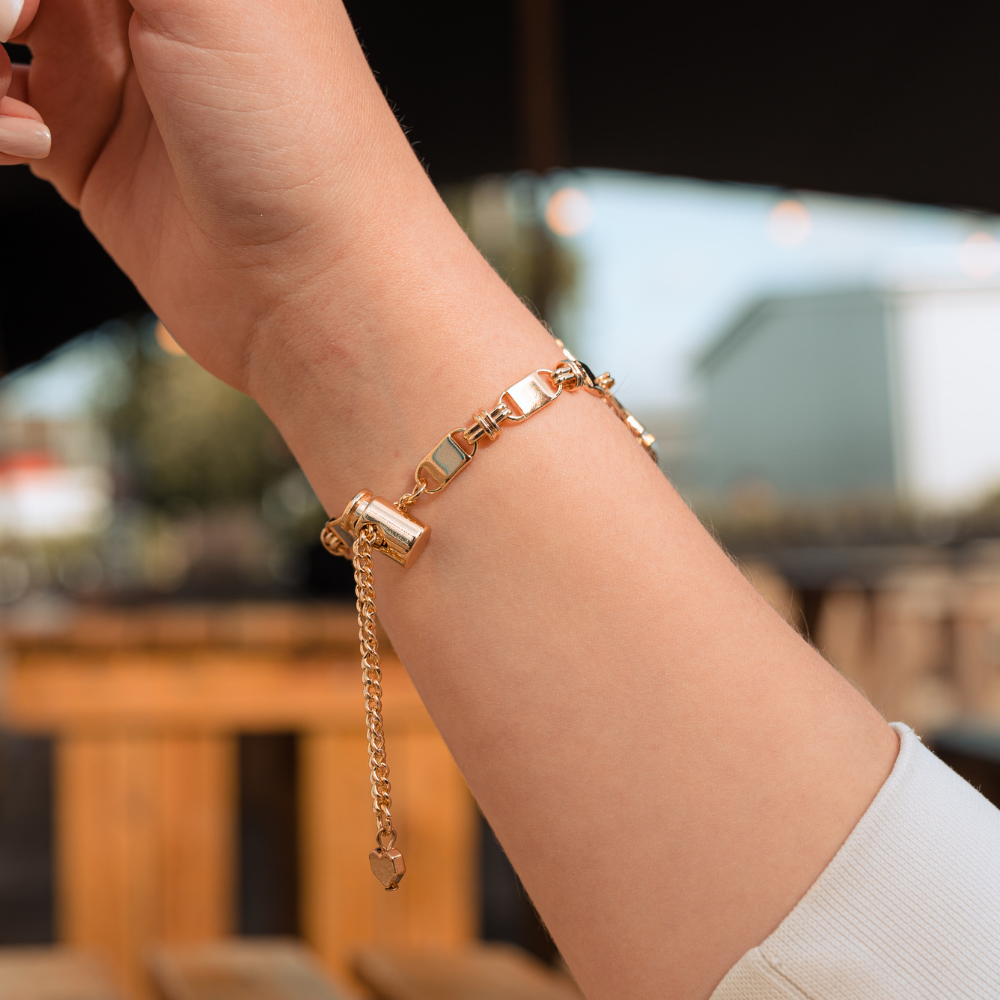 A close-up image of a person's wrist adorned with the Simple Gold Bracelet Apple Watch Strap by Coconut Lane. The strap features a cylindrical clasp and a chain with geometric links. The background is slightly blurred, showing outdoor seating and greenery. The person is wearing a light-colored sleeve.