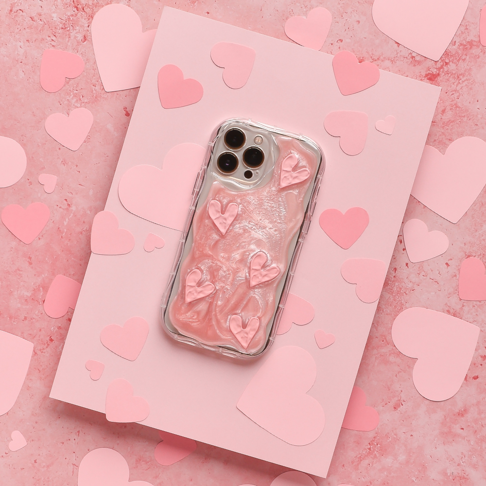 A smartphone encased in Coconut Lane's Shimmer Pink Hearts Phone Case is placed on a pink card surrounded by paper hearts. The sparkling pink hearts on the glossy, raised texture of the case stand out against the pink background adorned with various heart shapes.