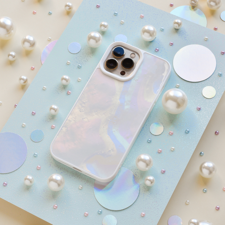 An iridescent iPhone with a Coconut Lane Mother of Pearl Phone Case is displayed on a light blue sparkly board. The background is adorned with various sizes of pearls and holographic circles, creating a whimsical and elegant arrangement.