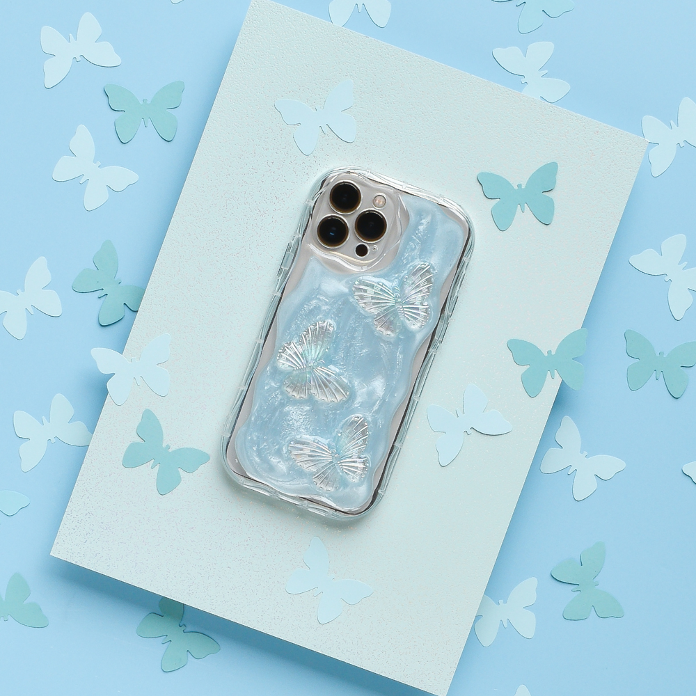 A smartphone with the Coconut Lane Shimmer Blue Butterflies Phone Case rests on a textured blue surface, surrounded by various paper cutouts of butterflies in shades of blue and white. The phone's camera lenses are visible at the top.