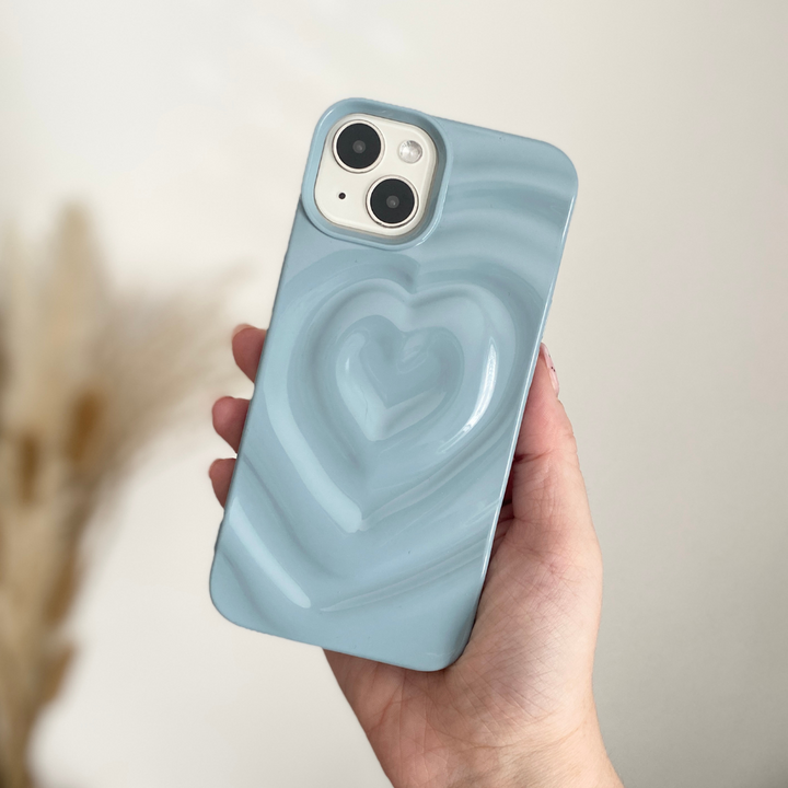 A hand holding Coconut Lane's Melting Heart Phone Case in Baby Blue, featuring a raised heart design on the back. The textured melting heart adds a unique touch, while the background is blurred with neutral tones.