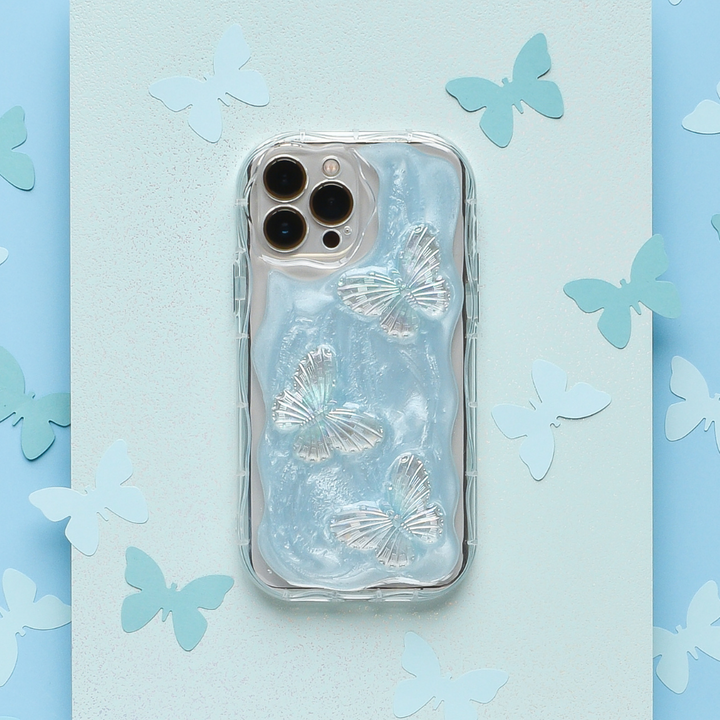 A silver smartphone fitted with Coconut Lane's Shimmer Blue Butterflies Phone Case, adorned with three iridescent butterfly designs, is surrounded by cut-out paper butterflies on a pastel blue and turquoise background. The phone features a triple camera on the top left corner.