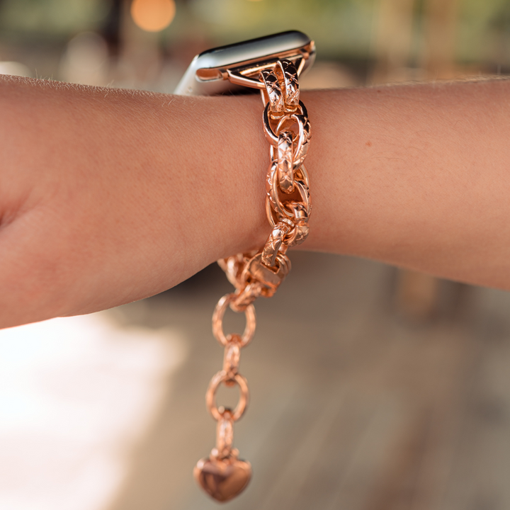 Close-up of a person's wrist showcasing the Chain Bracelet Apple Watch Strap - Rose Gold by Coconut Lane, featuring a heart-shaped charm. The person is stylishly pairing the rose gold strap with an Apple Watch, creating a cohesive and elegant look. The blurred background draws attention to these chic wrist accessories.