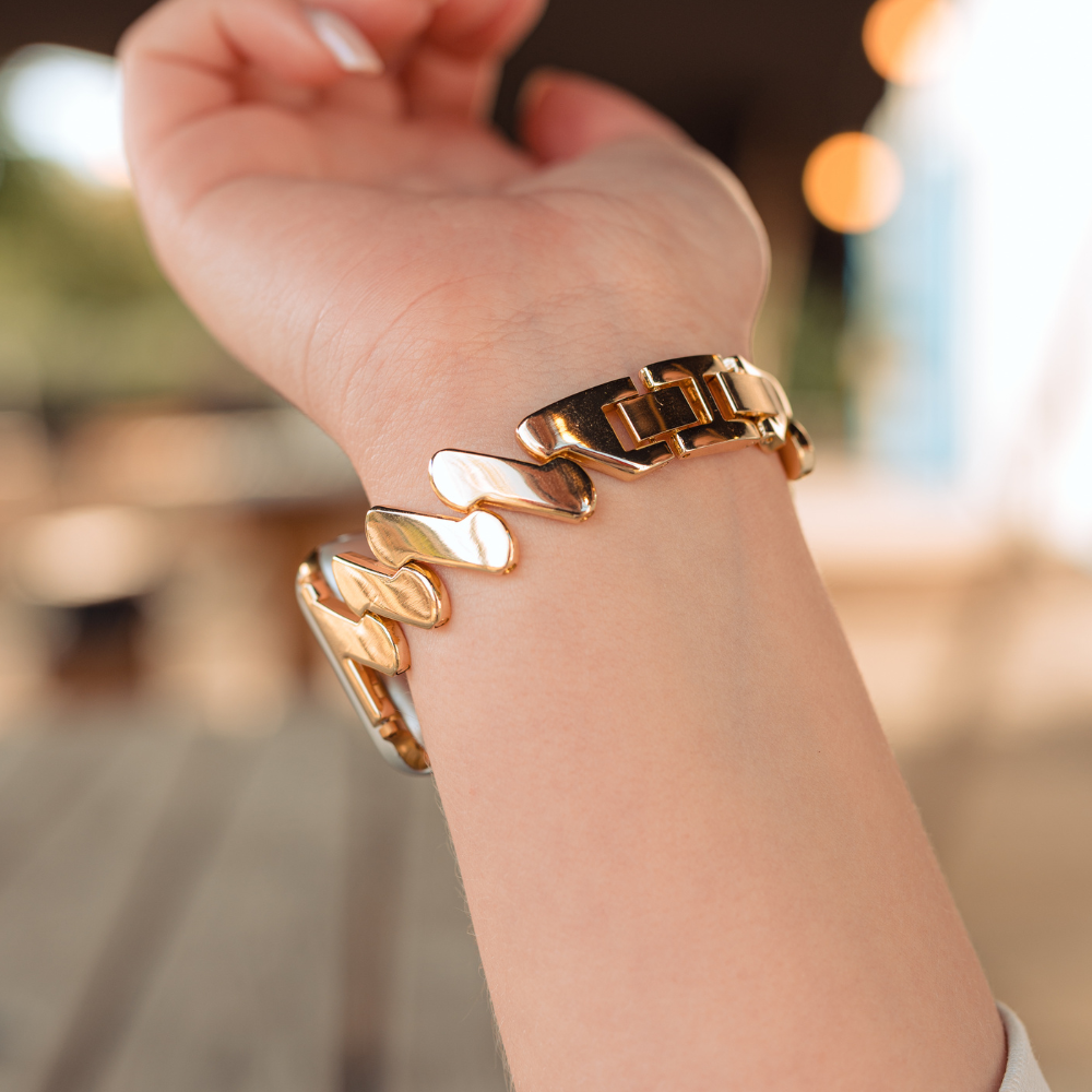 A close-up photo of a wrist wearing the Get Wavy Apple Watch Strap - Gold by Coconut Lane, showcasing its chunky wave-like and link elements. The background is slightly blurred, featuring an outdoor setting with a wooden table and blurry lights. The skin tone of the wrist is light.