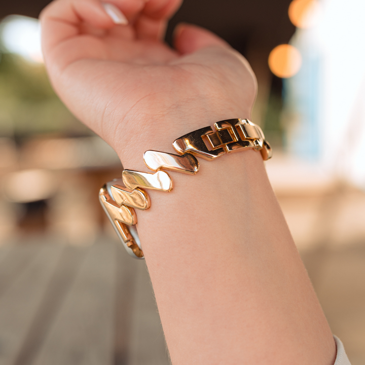 A close-up photo of a wrist wearing the Get Wavy Apple Watch Strap - Gold by Coconut Lane, showcasing its chunky wave-like and link elements. The background is slightly blurred, featuring an outdoor setting with a wooden table and blurry lights. The skin tone of the wrist is light.