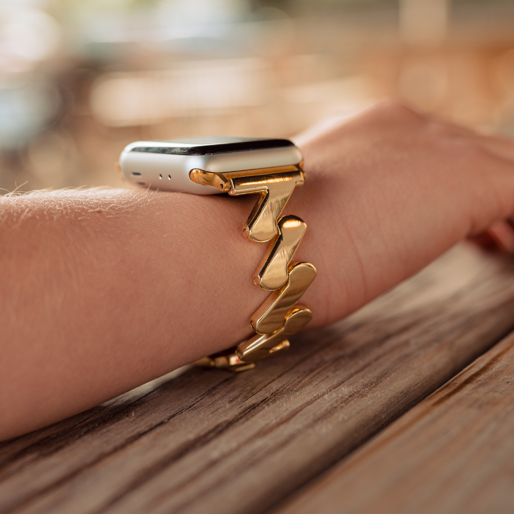 A close-up photo of a person's wrist wearing an Apple Watch with the Coconut Lane Get Wavy Apple Watch Strap in gold. The wrist and watch rest on a wooden surface, with the background softly blurred. The focus is on the elegant band, perfectly compatible with various Apple Watch models, and the sleek design of the smartwatch.
