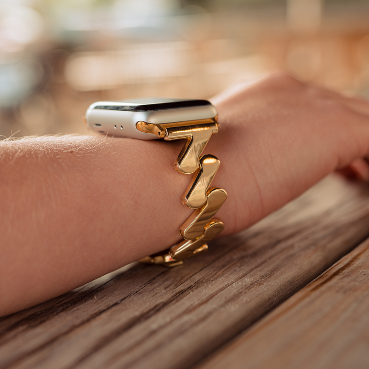 A close-up photo of a person's wrist wearing an Apple Watch with the Coconut Lane Get Wavy Apple Watch Strap in gold. The wrist and watch rest on a wooden surface, with the background softly blurred. The focus is on the elegant band, perfectly compatible with various Apple Watch models, and the sleek design of the smartwatch.