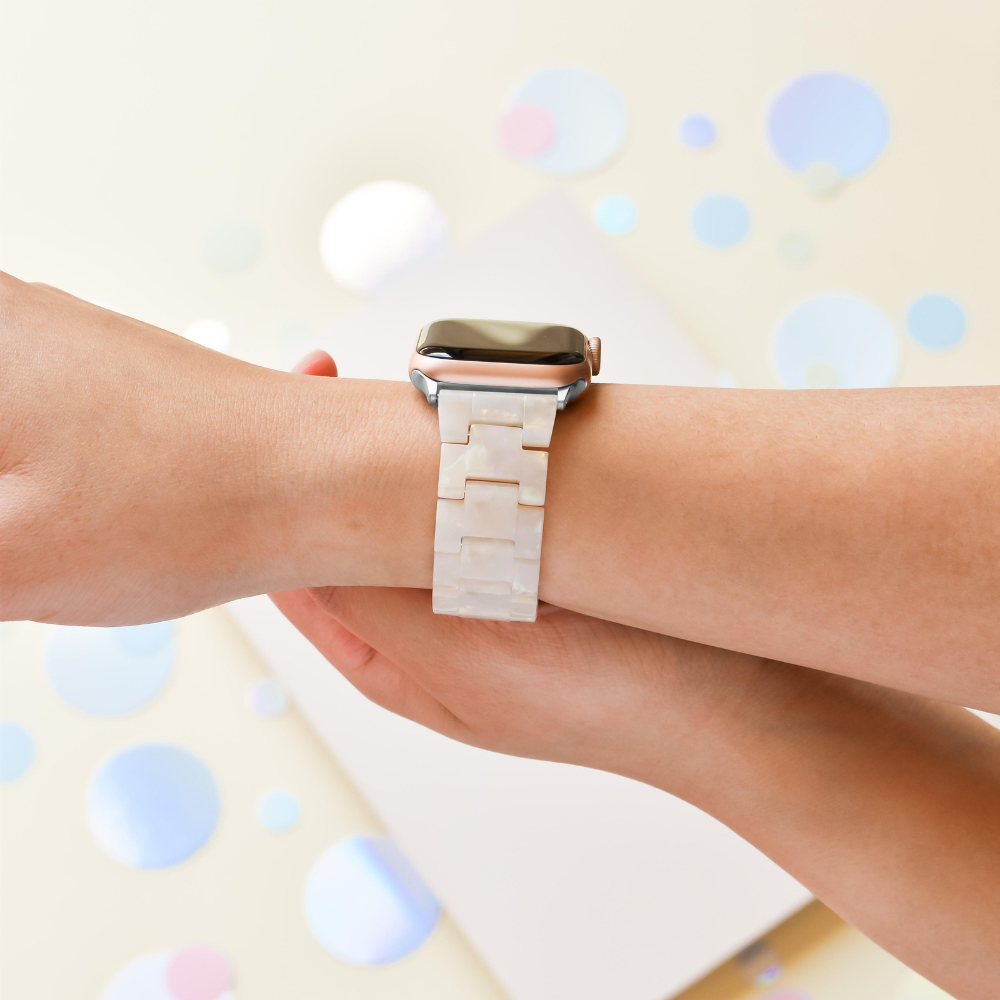 A person's wrist wearing a silver smartwatch featuring the Luxe Iridescent Apple Watch Strap by Coconut Lane, which has a white, marbled pattern. The background is a light cream color decorated with scattered iridescent circles.