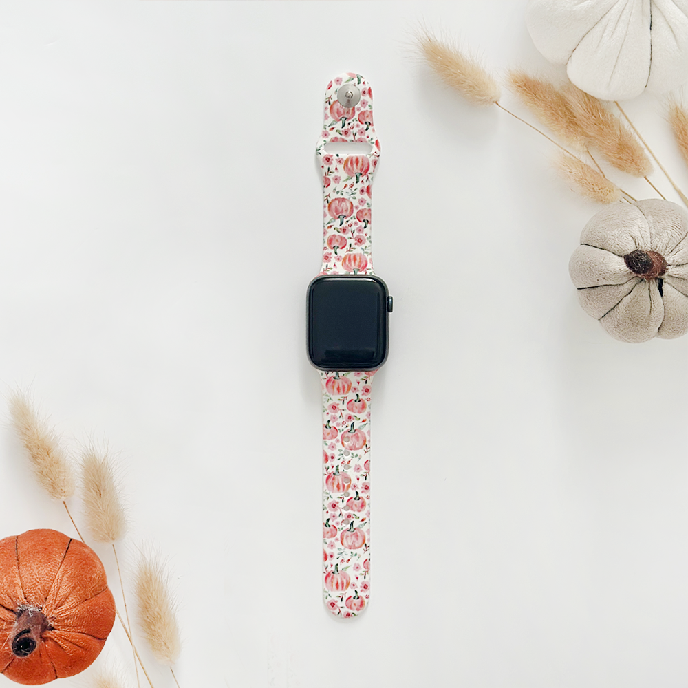 Coconut Lane Apple Watch strap with ditsy pumpkin pattern, shown beside fabric pumpkins and dried grasses on white background.