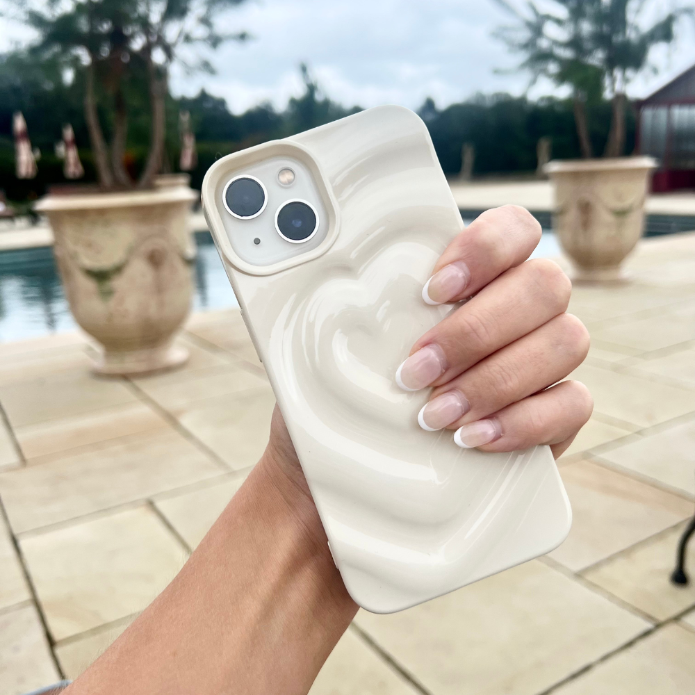 A hand with manicured nails holds the Melting Heart Phone Case - Coconut Cream by Coconut Lane, featuring a cream-colored, heart-patterned design. The background showcases an outdoor area with large potted plants, a swimming pool, and trees in the distance.